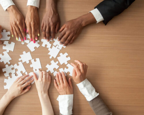 Hands of multi-ethnic team assembling jigsaw puzzle, multiracial group of black and white people joining pieces at desk, successful teamwork concept, help and support in business, close up top view practicing networking best practices